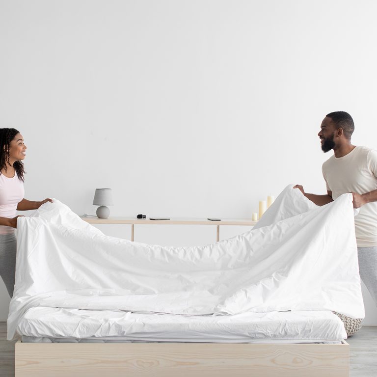 Cheerful young black woman and guy make bed and hold blanket in bedroom interior on white wall background in morning. Fun and household chores, cleaning at home together during covid-19, copy space